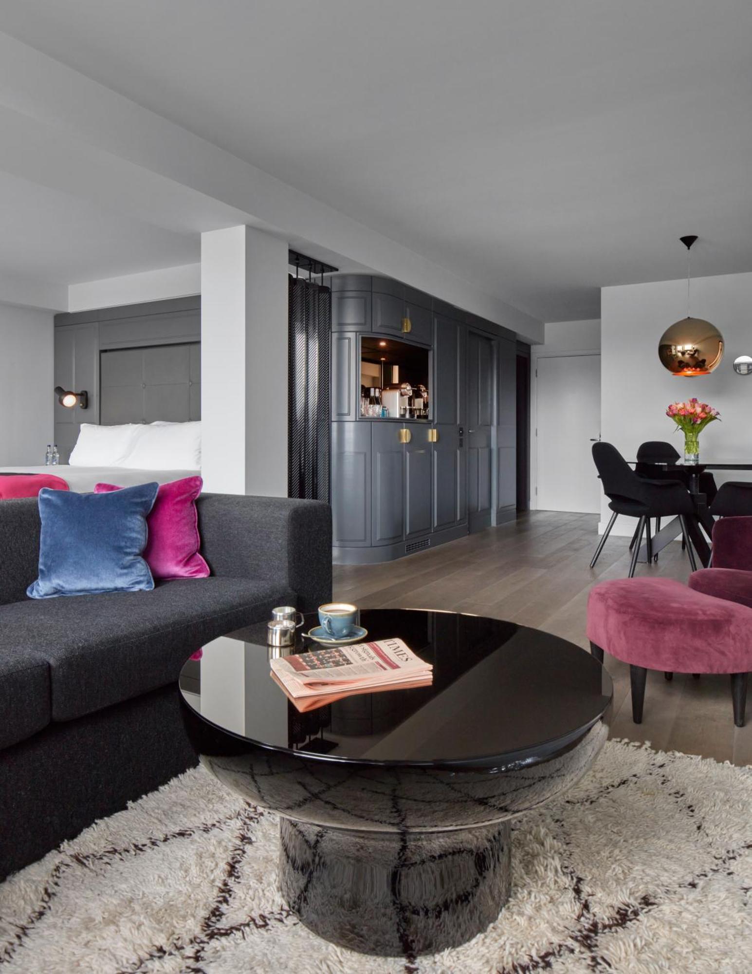 Hotel Sea Containers Londres Exterior foto The image shows a modern-looking living space, likely a hotel suite or stylish apartment. It features a dark gray sofa with colorful cushions in shades of blue and pink. In front of the sofa, there's a round black coffee table that holds a magazine a