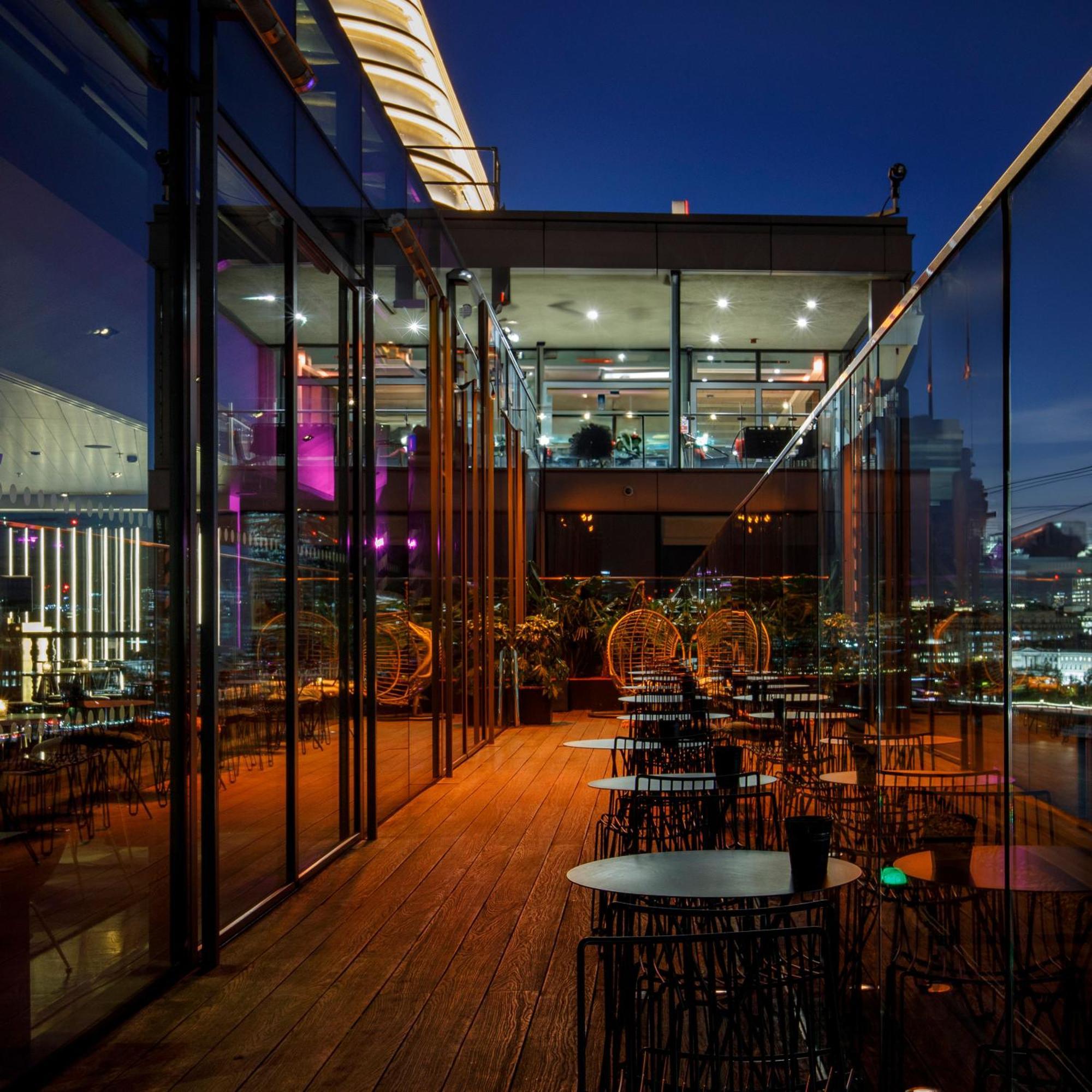 Hotel Sea Containers Londres Exterior foto The photo depicts a stylish restaurant or lounge area, featuring a modern design with extensive glass walls. It appears to be set on an upper floor, providing a view of the city skyline. The space has a wooden deck and several circular tables surroun